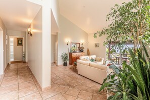 A spacious living area with a beige sofa, a wooden cabinet, and a dining table with chairs. There are potted plants and a large tree in the corner. - Casa Jardim Botanico (With Award Winning Garden) (Madeira)