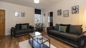 Ein Wohnzimmer mit zwei dunklen Sofas, einem Glastisch und einem Esstisch mit Stühlen. An den Wänden hängen Bilderrahmen, und links befindet sich eine Holztür.