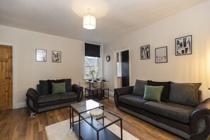Ein Wohnzimmer mit zwei dunklen Sofas, einem Glastisch und einem Esstisch mit Stühlen. An den Wänden hängen Bilderrahmen, und links befindet sich eine Holztür.