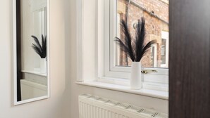 Ein Fenster mit einem weißen Heizkörper darunter, eine weiße Vase mit schwarzen Dekoelementen auf der Fensterbank und ein Spiegelbild darin.
