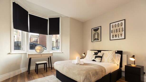 Ein Schlafzimmer mit einem Bett, Nachttischen, einem Schreibtisch, einem Fenster mit schwarzen Vorhängen und zwei Wandbildern.