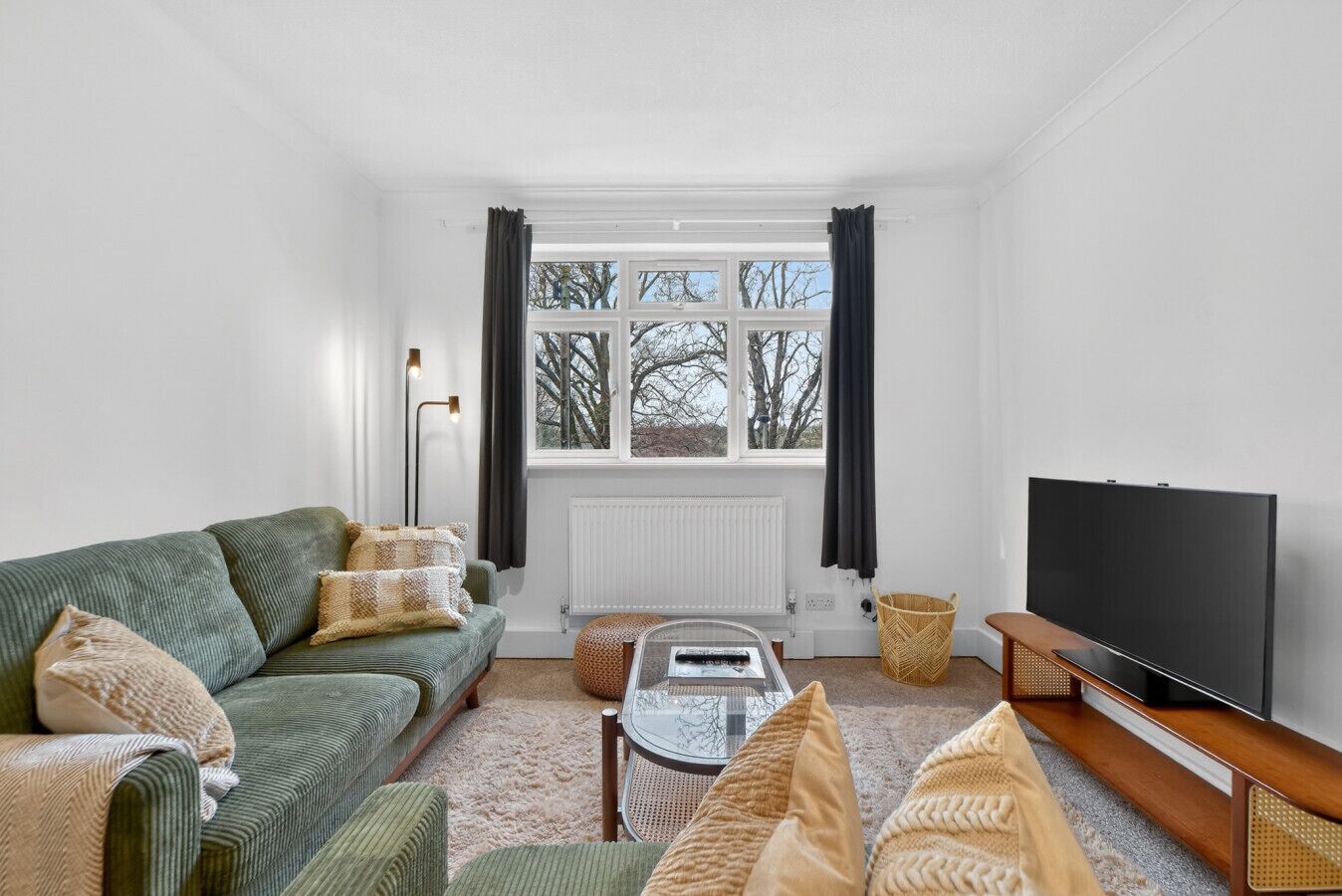 Een woonkamer met een groene bank, een glazen salontafel, een televisie en uitzicht op bomen door grote ramen.