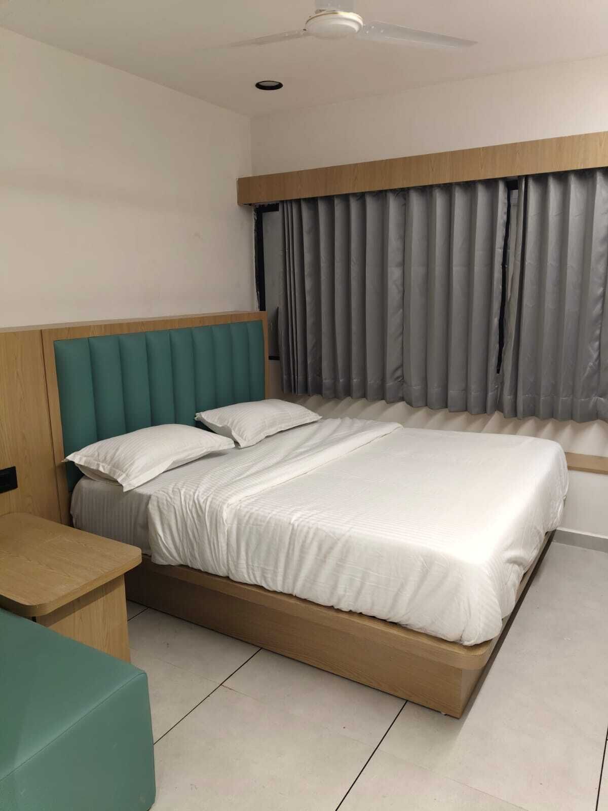Une chambre moderne avec un grand lit, une tête de lit en bois, une table de chevet et une fenêtre agrémentée de rideaux gris.