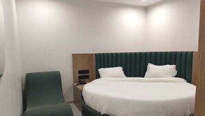A modern hotel room with a round bed, a green upholstered chair, a wooden nightstand, and a small table. - hotel shiva inn (Ahmedabad)