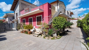 Una moderna casa a due piani con facciata rossa, un balcone e un piccolo giardino con piante in vaso e una palma.