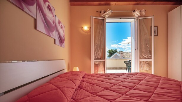 Camera da letto con un letto grande, un comodino, una lampada a muro e vista all'esterno tramite una porta aperta.