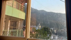 A room with a large window overlooking a mountainous area and a building with a green facade. - Hotel Laxman Grand (Rishikesh)
