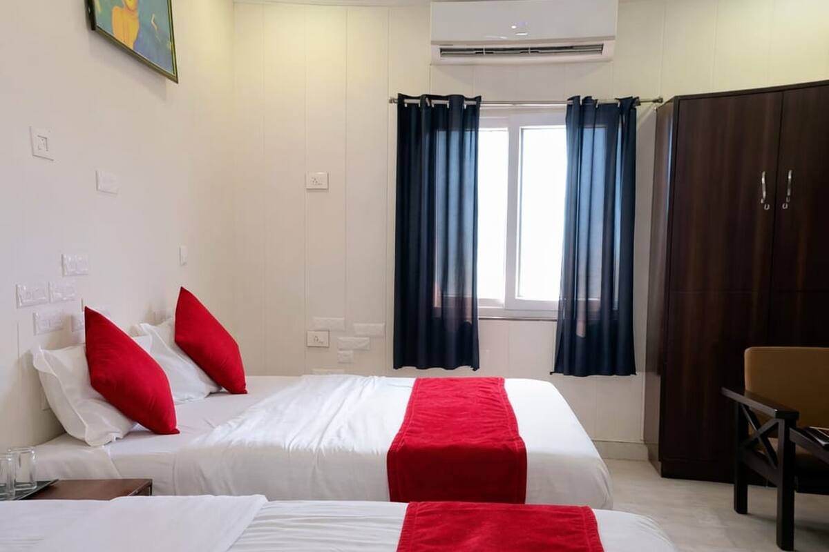 Habitación de hotel con dos camas, ropa de cama roja y blanca, ventilador de techo, unidad de aire acondicionado y ventana con cortinas.