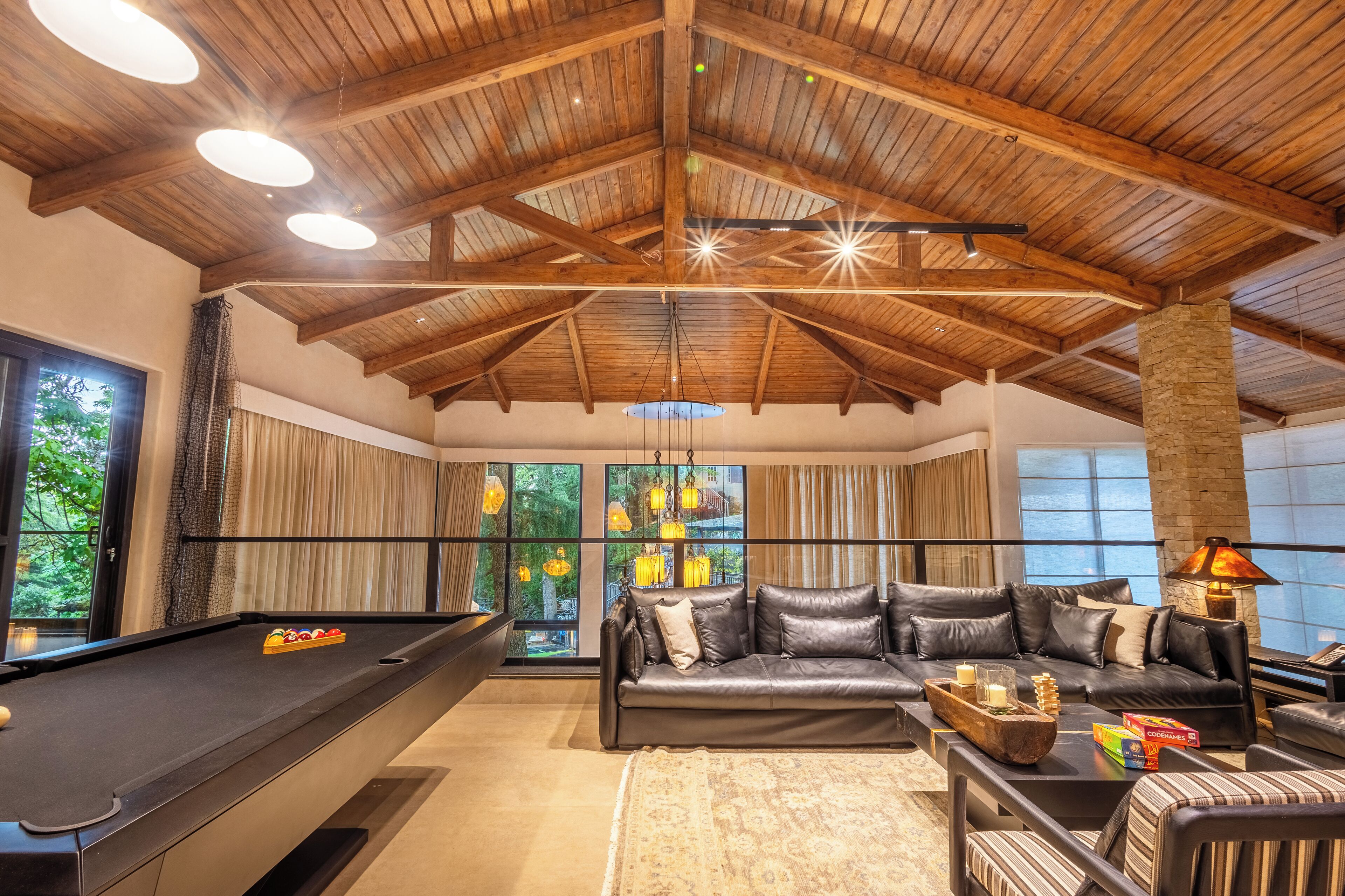 A spacious room with a pool table, leather sofa, and large windows offering a view of greenery.