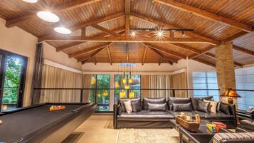 A spacious room with a pool table, leather sofa, and large windows offering a view of greenery.