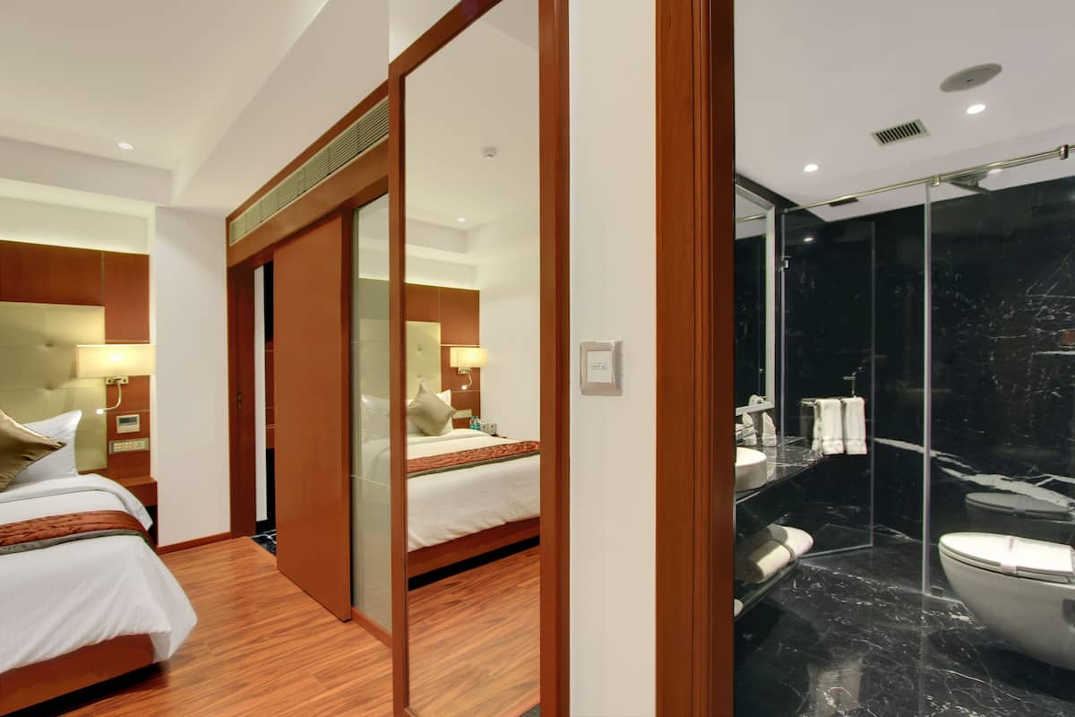 Habitación de hotel con piso de madera, cama con ropa blanca, un espejo grande, baño con piso de mármol negro y cabina de ducha de vidrio.