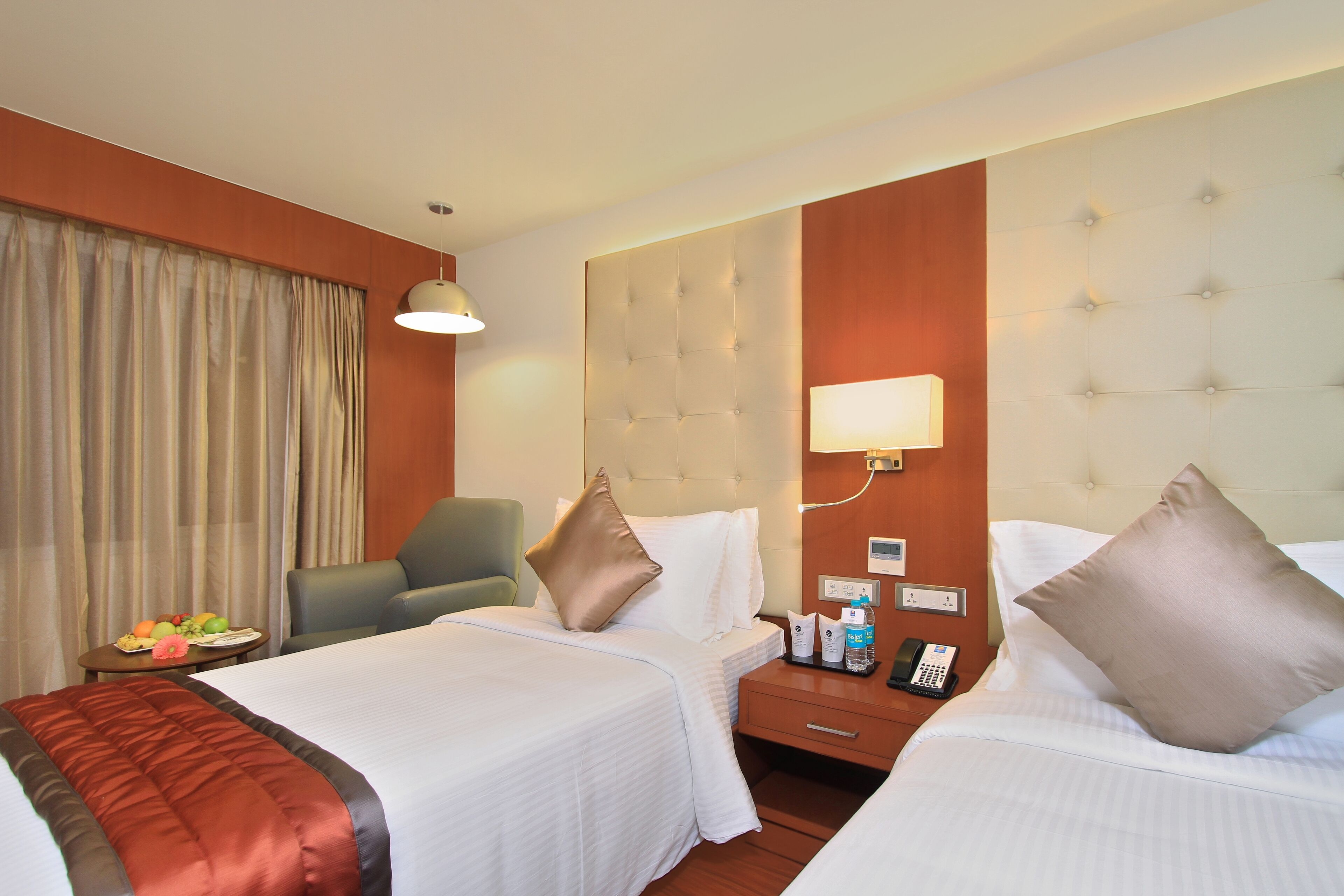 Habitación de hotel con dos camas, una mesita de noche con teléfono, lámpara y una cesta de frutas sobre una pequeña mesa.