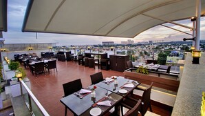 An outdoor dining area with tables and chairs, a large umbrella, and a city view in the background. - Lavender Premier (Bengaluru)