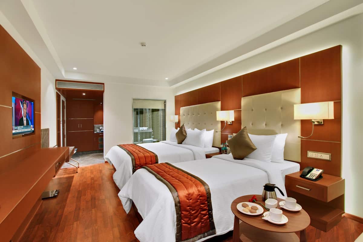 Habitación de hotel con dos camas, un televisor, una mesa de centro con provisiones para el desayuno y un baño visible a través de una puerta abierta.