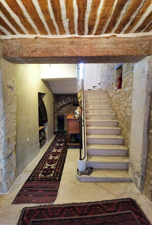 Un escalier étroit aux murs de pierre et au plafond en bois. Un tapis à motifs recouvre les marches, et une petite armoire en bois est visible au bas de l’escalier.