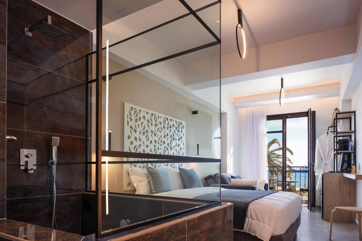 A modern hotel room with a large bed, a glass shower enclosure, a balcony with a view of the sea, and a wooden desk.