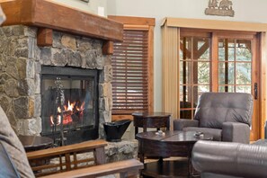 A stone fireplace with a fire, a wooden bench, a leather armchair, and a large window with blinds.