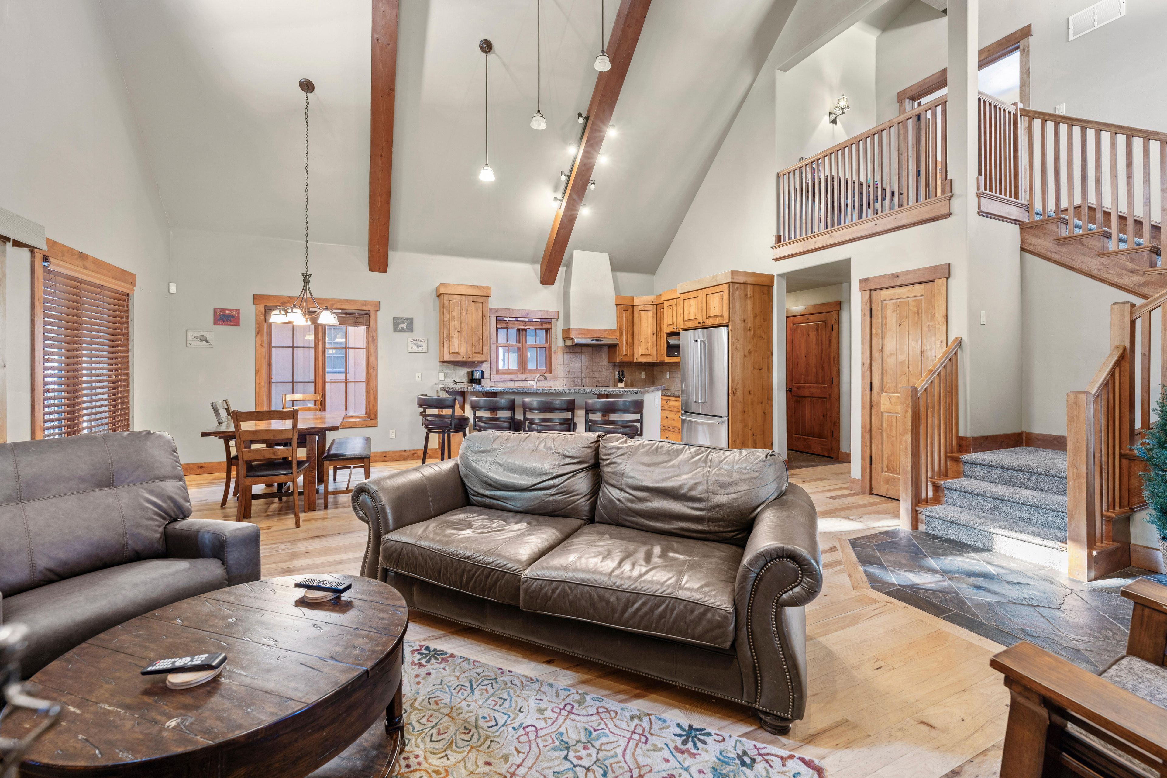 A spacious living area with a leather sofa, wooden coffee table, and a dining table with chairs. A kitchen with wooden cabinets and a refrigerator is visible in the background.