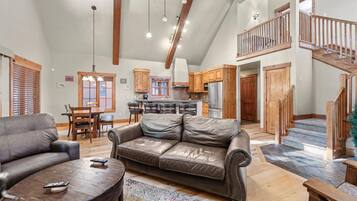 A spacious living area with a leather sofa, wooden coffee table, and a dining table with chairs. A kitchen with wooden cabinets and a refrigerator is visible in the background.