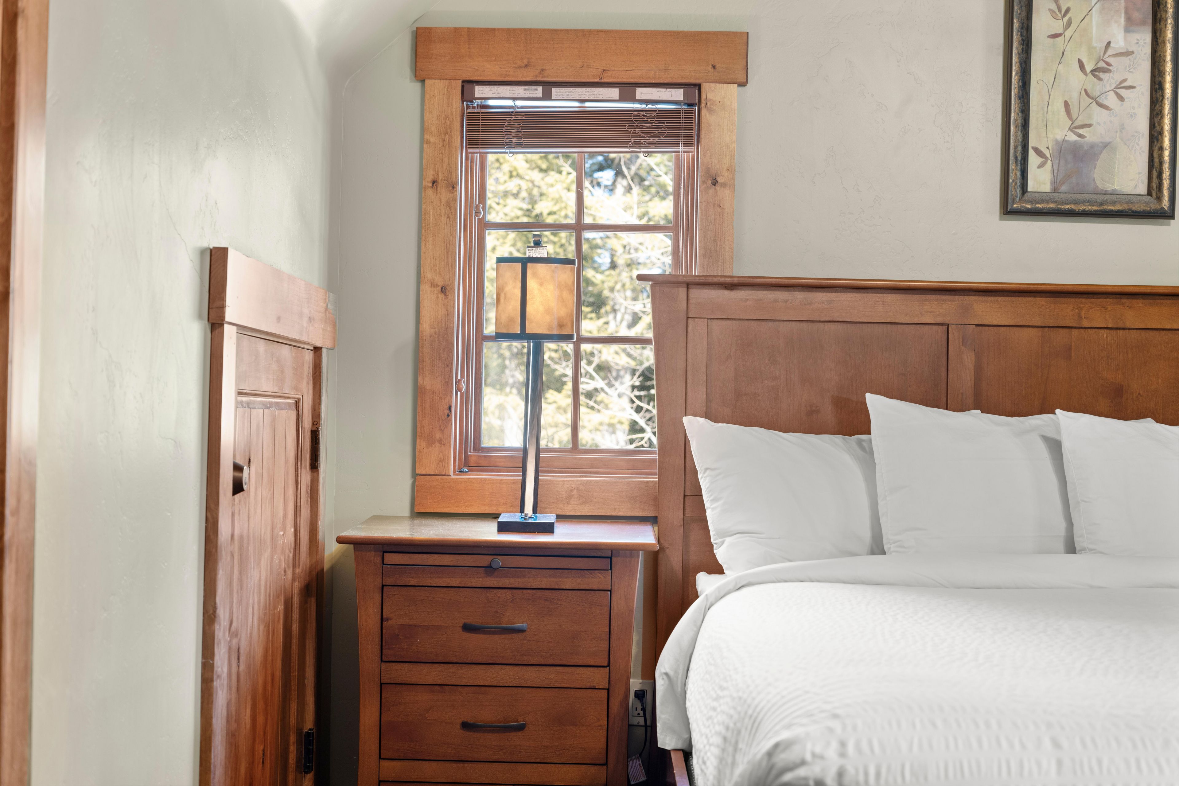 A bedroom with a wooden headboard, a bedside table with drawers, a lamp, and a window with blinds.