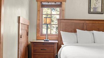 A bedroom with a wooden headboard, a bedside table with drawers, a lamp, and a window with blinds.