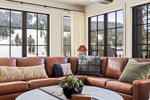A brown leather sofa with multiple cushions, a small table with a plant, and large windows with a snowy mountain view. - Alpine Glow Retreat | Exclusive Tamarack Perks (Tamarack)