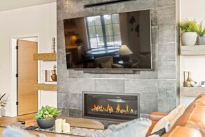 A modern living room with a flat-screen TV mounted on a stone wall, a fireplace with a visible fire, and a cozy seating area with a leather couch. - Alpine Glow Retreat | Exclusive Tamarack Perks (Tamarack)