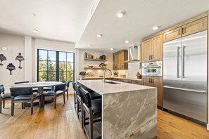 A modern kitchen with a central island, stainless steel appliances, and a dining area with a view. - Alpine Glow Retreat | Exclusive Tamarack Perks (Tamarack)