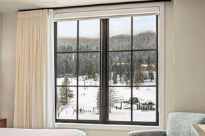 A hotel room with a large window offering a view of a snowy landscape with trees and a ski lift. - Alpine Glow Retreat | Exclusive Tamarack Perks (Tamarack)