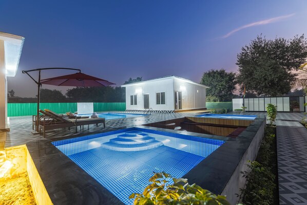 A swimming pool area with lounge chairs and a small building, likely a spa or sauna, in the background. - Alaya Stays Krishna Farm With Pool (Noida)