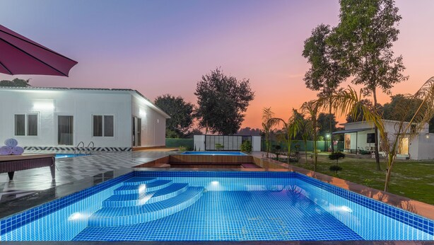En swimmingpool med blå fliser, en bygning med hvide vinduer og en solnedgang i baggrunden.