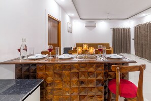 A dining area with a wooden table set for two, a sofa in the background, and a red chair in the foreground. - Alaya Stays Krishna Farm With Pool (Noida)