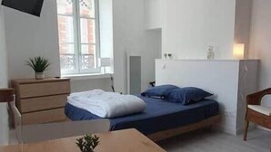 A modern bedroom with a bed, a bedside table, a chair, a TV mounted on the wall, and a window with a view of buildings outside.