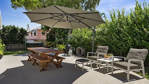 Um pátio externo com mesa e bancos de madeira, um guarda-sol bege e um espaço de estar com almofadas.