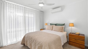 Quarto com cama, duas mesas de cabeceira, ventilador de teto e ar condicionado fixado na parede.