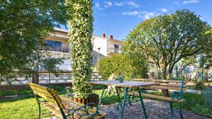 Zona d'estar a l'aire lliure amb una taula i una cadira de fusta, envoltada de vegetació i un edifici al fons.