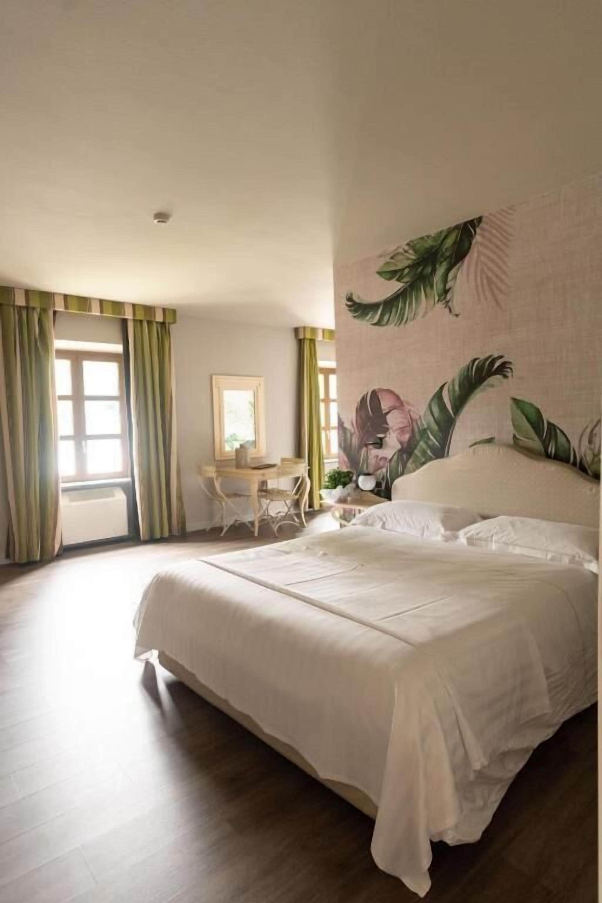 Un dormitorio con una cama grande, un escritorio y una silla. Hay una ventana con cortinas y una pared con un diseño de hojas.