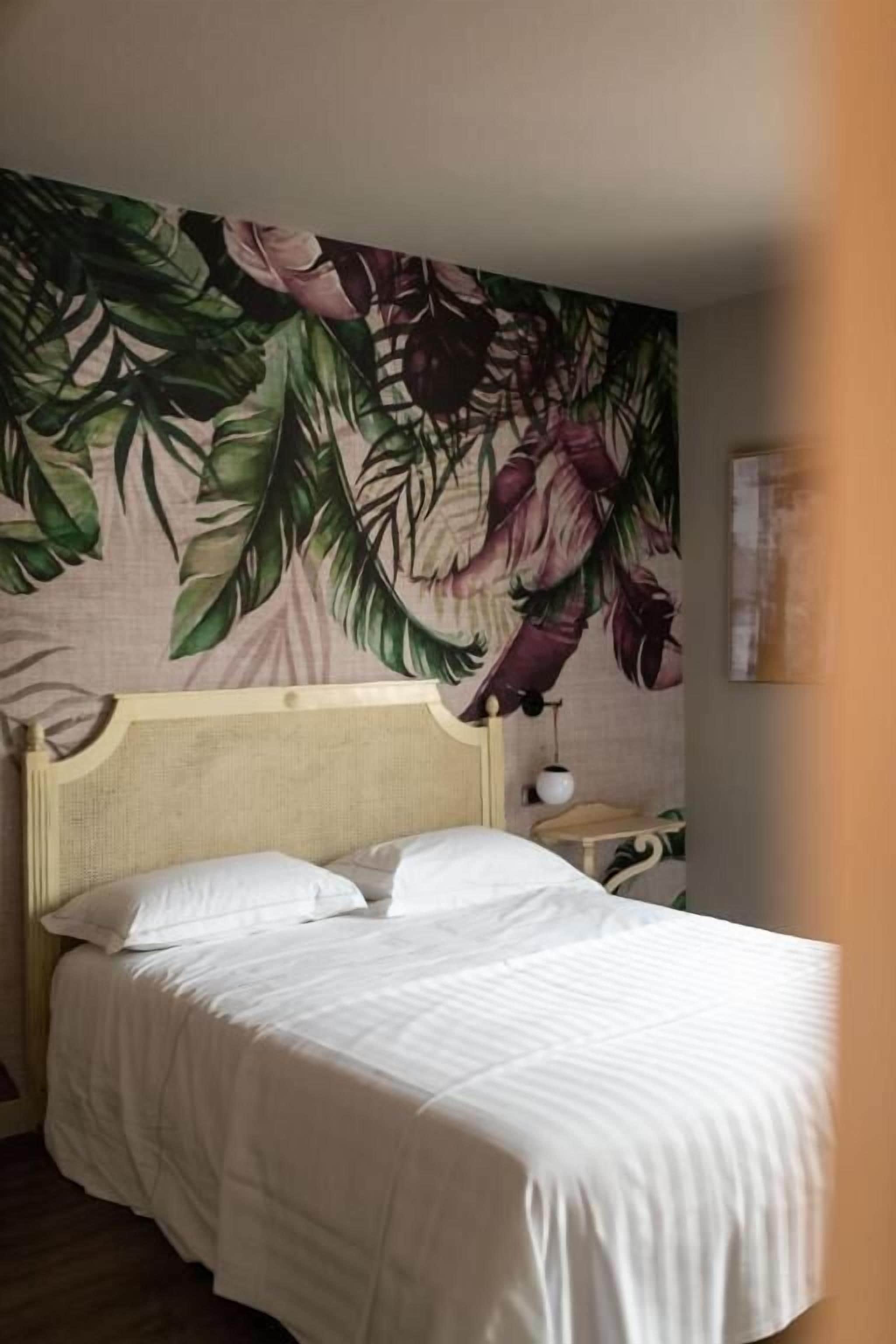 Un lit bien fait, avec une tête de lit, un mur orné d’un motif de feuilles tropicales et une petite table de chevet avec une lampe.