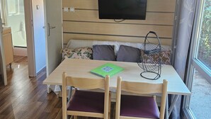 A modern living room with a wooden table, two chairs, a sofa with cushions, a mounted TV, and a window with curtains. - Quiet chalet under the trees 3 km from Bonifacio and 2 km from the beaches (Bonifacio)