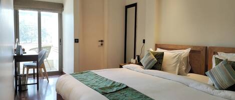 A hotel room with a large bed, a desk with a chair, a mirror, and a view through a door.