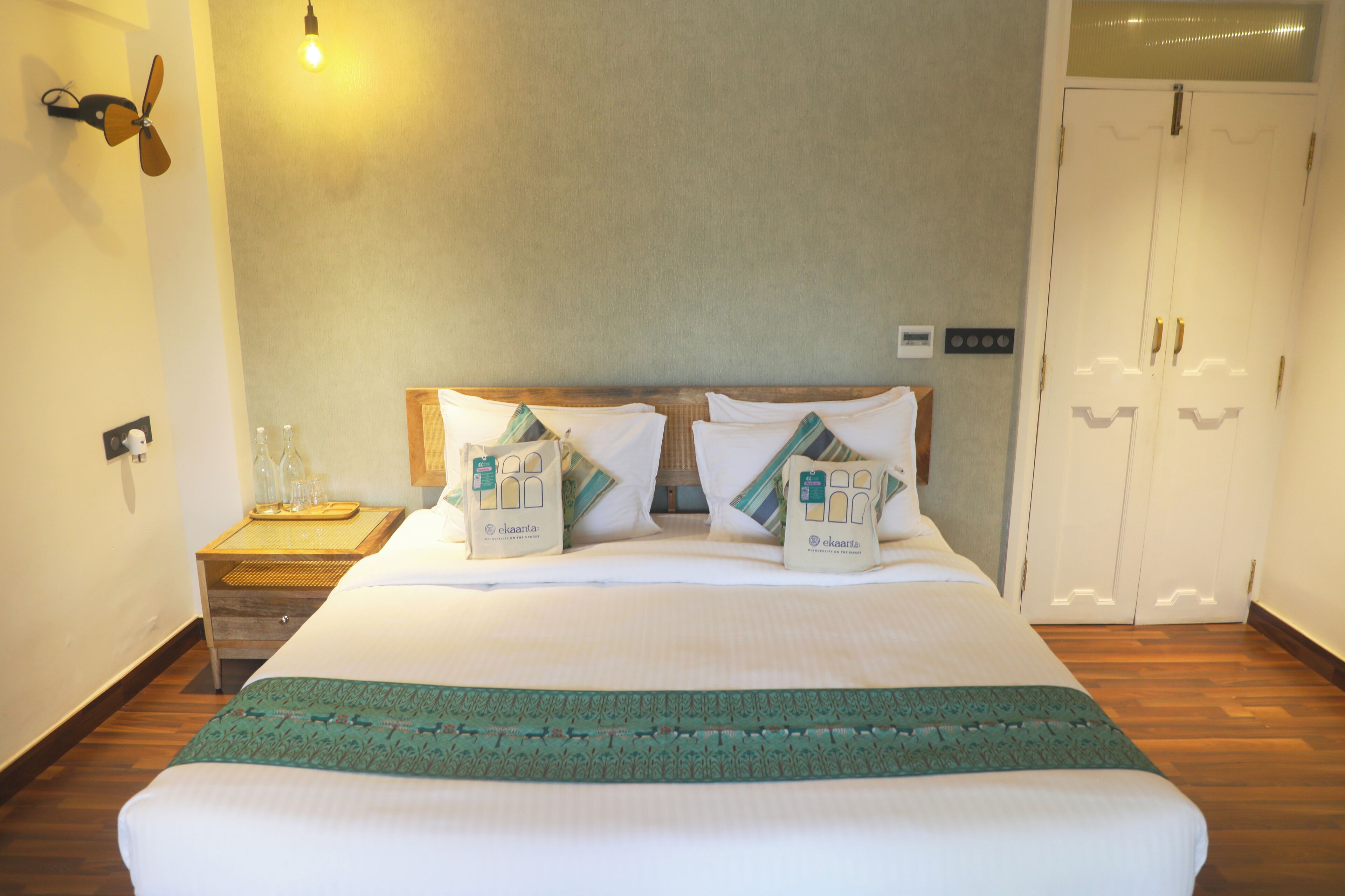 A neatly made bed with a green bedspread, two pillows with a geometric pattern, a bedside table with a tray, and a wall-mounted light fixture.