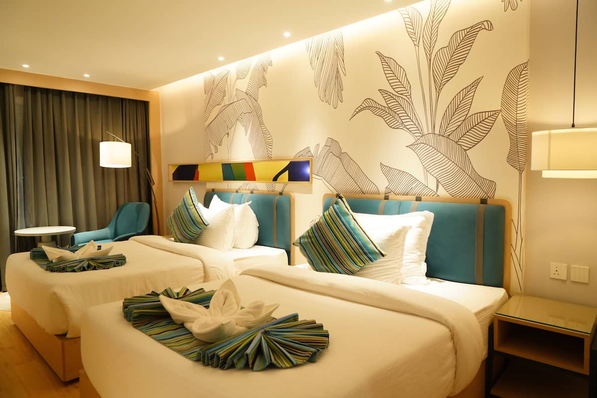 Quarto de hotel com duas camas, uma escrivaninha, uma cadeira e uma parede com padrão de folha.