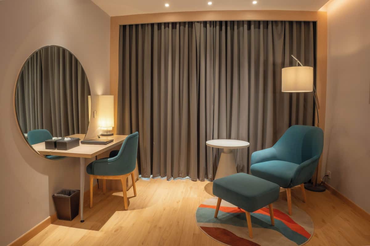Quarto de hotel com um espelho redondo, uma secretária com candeeiro, uma cadeira azul, uma mesa redonda pequena e uma janela grande com cortinas.