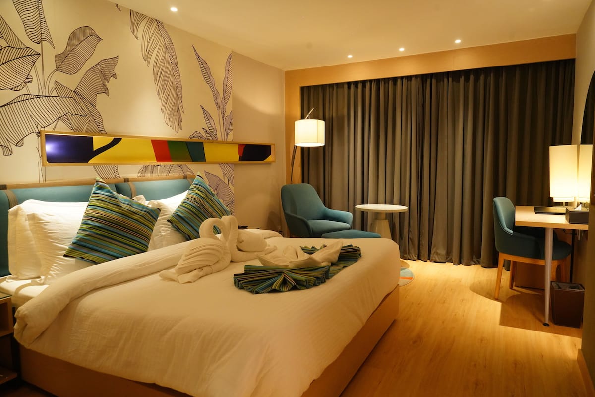 Quarto de hotel com cama, escrivaninha, cadeira, abajur e cortina.