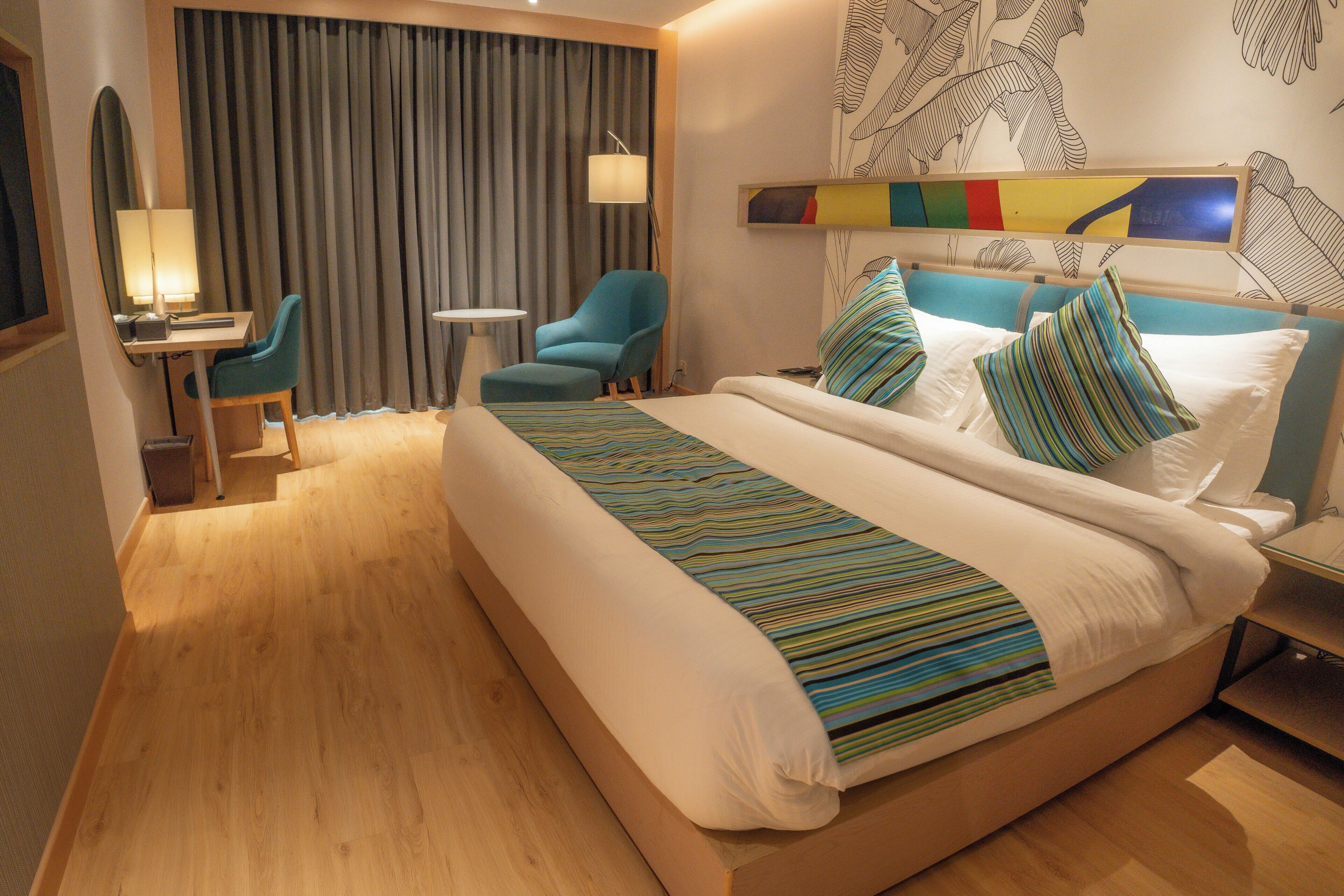 A hotel room with a large bed, a desk with a chair, a small table, and a wall-mounted lamp.