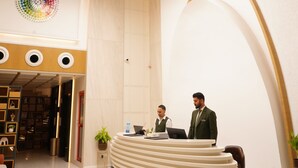 A reception desk with two staff members, a modern chandelier, and a large circular logo on the wall.