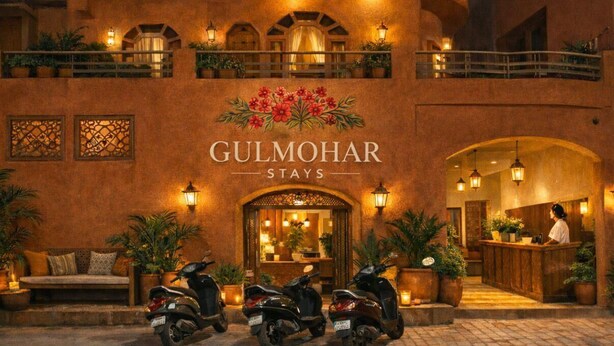 A building with a sign reading "Gulmohar Stays" and a reception area with a receptionist. There are potted plants and a scooter parked in front. - Gulmohar Stays (siddhagiribagh)