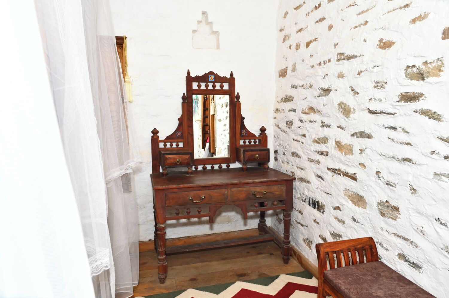 A wooden dresser with drawers and a mirror, a chair, a patterned rug, and a white brick wall.