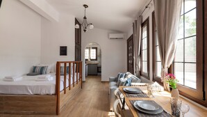 Una habitación amplia con piso de madera, una cama, un sofá, una mesa de comedor lista para dos personas y ventanas grandes con cortinas.
