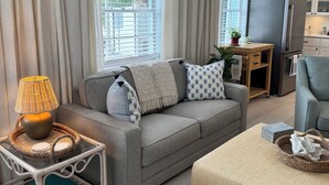 Een moderne woonkamer met een grijze bank, een klein bijzettafeltje met een lamp, een salontafel en een keukenhoek met een koelkast en kasten.